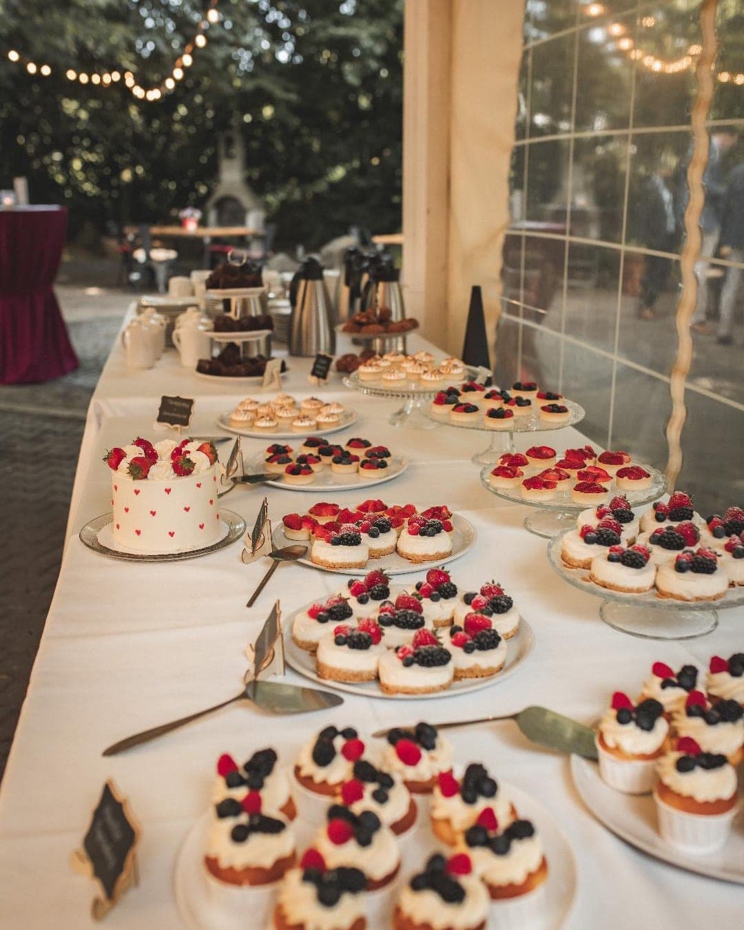 Sweet Table auf einer langen weiß gedeckten Tafel: eine kleine runde Torte mit Erdbeeren und roten Herzen, dazu Käsekuchen-Törtchen und Cupcakes mit Himbeeren, Brombeeren und Heidelbeeren sowie Baiser-Tartelettes